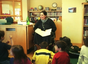Sharon in an elementary school library, a folder of papers in her hand, wearing an oxygen canula, leaning forward with her mouth open, as if reading or talking. Gadget lies on the ground next to her in a green pack, looking up at her. In the foreground are several first-graders, looking in many different directions, some of them obviously moving around.