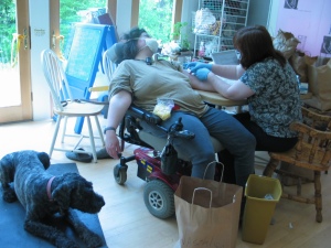Barnum lies on a black yoga mat next to Sharon, who is in her powerchair with the back reclined all the way and her feet slid off the footplate. She's wearing a large white mask covering all of her face but from her eyes up. One arm is hanging over the side of the armrest, the other is outstretched on the table where a nurse in a medical mask and gloves is changing Sharon's PICC line dressing.