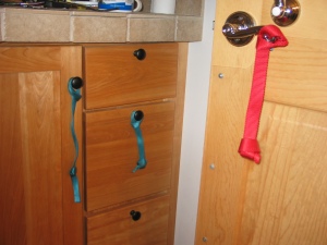 A door with a metal door lever with a red nylon webbing pull attached. It has a knot in the bottom. Next to the door is a cupboard, with a cabinet door and three drawers. Thin, turquoise nylon pulls hang from the cabinet doorknob and the knob of one of the drawers.