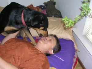 Marlena laying on her back in bed. A black-and-tan short-haired large-breed older puppy -- maybe part Doberman or Rottweiler -- wearing a pink collar is licking her from her chin to her eyebrow. Marlena's eyes are closed and her hand rests on the dog's chest.