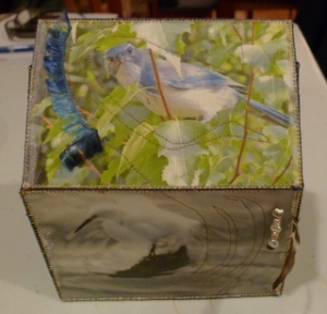 A fabric-covered box. The top shows a blue jay in a green leafy tree with a blue feather attached to it, and the side shows a gorgeous white ibis about to take off over stormy waters.