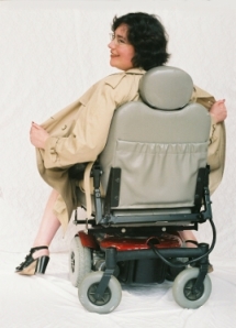 Sharon sits in her powerchair, which is backwards; both Sharon and the chair are facing away from the camera. The chair has a gray seat and wheels, with a cherry-red base. Sharon wears a tan trench-coat, and she is holding it open on both sides, as if she is flashing whoever is in front of her. All that can be seen of Sharon are her naked calves, her feet in four-inch-high patent-leather heels, her hands pulling open the trench-coat, and her head. She is turning her head toward the camera, so her face is in profile, smiling mischievously. Her facial expression is saucy, letting you know that she does not take herself too seriously.