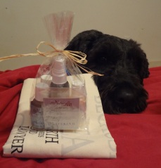 Clear cellophane bag tied with a straw bow with many small bottles in it, sitting on a folded tote bag. Barnum rests his chin on the bed just behind the items.